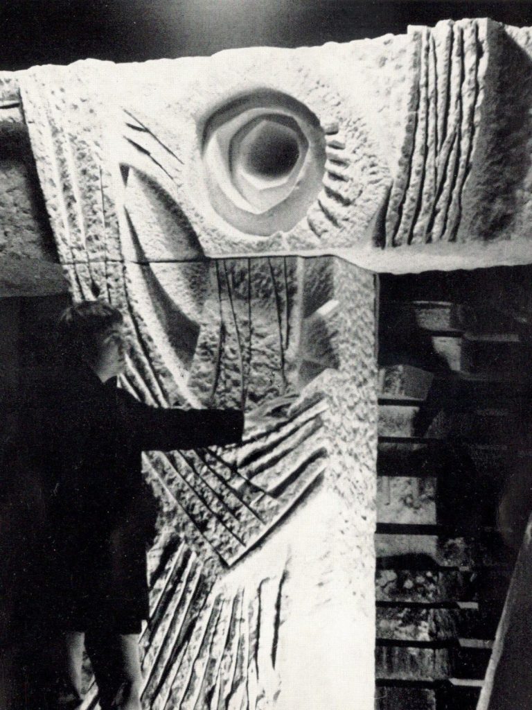 Lebensbaum Eine große Steinskulptur in T-Form, vor der ein Mensch steht